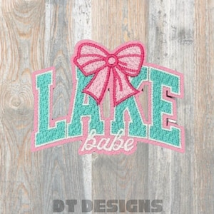 May include: A pink and teal embroidered patch with the words "LAKE babe" in a collegiate font, topped with a pink bow. The patch is set against a wooden background, with the text "DT DESIGNS" at the bottom.