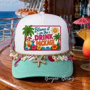 May include: A white and teal trucker hat featuring a rectangular patch with the text Blame it on the DRINK PACKAGE, palm trees, a sun, and tropical flowers. The brim is adorned with a gold chain, white pearls, and various sea shell charms.