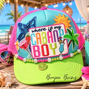 May include: A light blue and lime green trucker hat featuring a central patch with the text "where if my CABANA BOY" surrounded by embroidered icons of a sun, a palm tree, and cocktails. The hat is accented with a beaded trim and a lime slice charm.