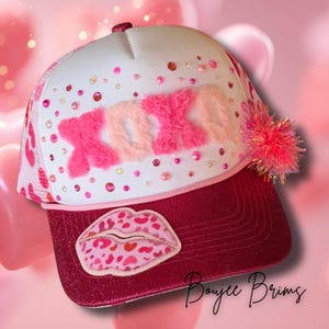 May include: A pink and white trucker hat with the word "XOXO" in fluffy pink letters. The hat features a glittery red brim and a lip-shaped patch. Embellished with rhinestones and a pink pom-pom.