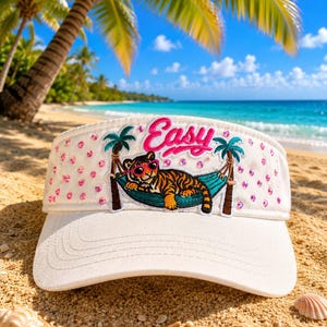 May include: A white visor featuring an embroidered patch of a tiger wearing pink sunglasses while resting in a teal hammock between two palm trees. The word Easy is embroidered in pink above the tiger, and the visor is decorated with pink sequins.
