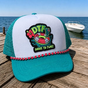 May include: A trucker hat with a white front panel and a teal mesh back. The hat features a patch with the text "DTF" above a graphic of a life preserver and a cocktail. A line of pink rhinestones adorns the brim.