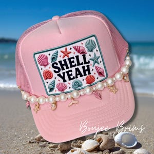 May include: Pink trucker hat with a mesh back and a decorative pearl chain. The front patch reads "SHELL YEAH" with seashell and starfish graphics. The hat is embellished with charms and is on a sandy beach.