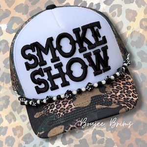May include: A trucker hat with a white front panel and a camouflage and leopard print brim. The hat features the words "SMOKE SHOW" in large, black, embroidered letters. A beaded chain with dice accents the brim.