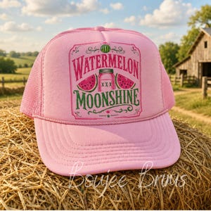 May include: A light pink trucker hat featuring a graphic print of a mason jar, watermelon slices, and the text Watermelon Moonshine. The cap has a structured front panel, a curved brim, and a mesh back with a plastic snapback closure for adjustment.