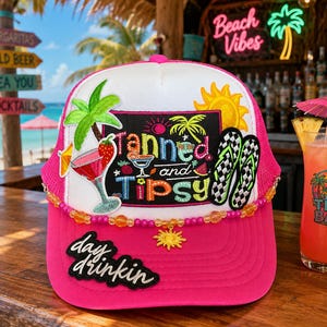 May include: A bright pink and white trucker hat featuring a central patch with the text Tanned and Tipsy, a palm tree, a cocktail, a sun, and checkered flip-flops. The hat is adorned with a beaded trim and a sun charm, plus a day drinkin patch.