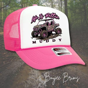 May include: A pink and white trucker hat with a curved brim. The hat features a graphic of a pink and leopard print off-road vehicle with the text "Life Is Better Muddy". The hat has a pink mesh back.