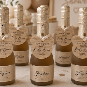 Puede incluir: Seis botellas miniatura de vino espumoso Freixenet con tapas y etiquetas de papel aluminio dorado. Cada botella tiene una etiqueta de madera clara en forma de ratón atada con una cinta blanca, con el texto: POP & CHEER WHEN Baby Brown IS HERE.