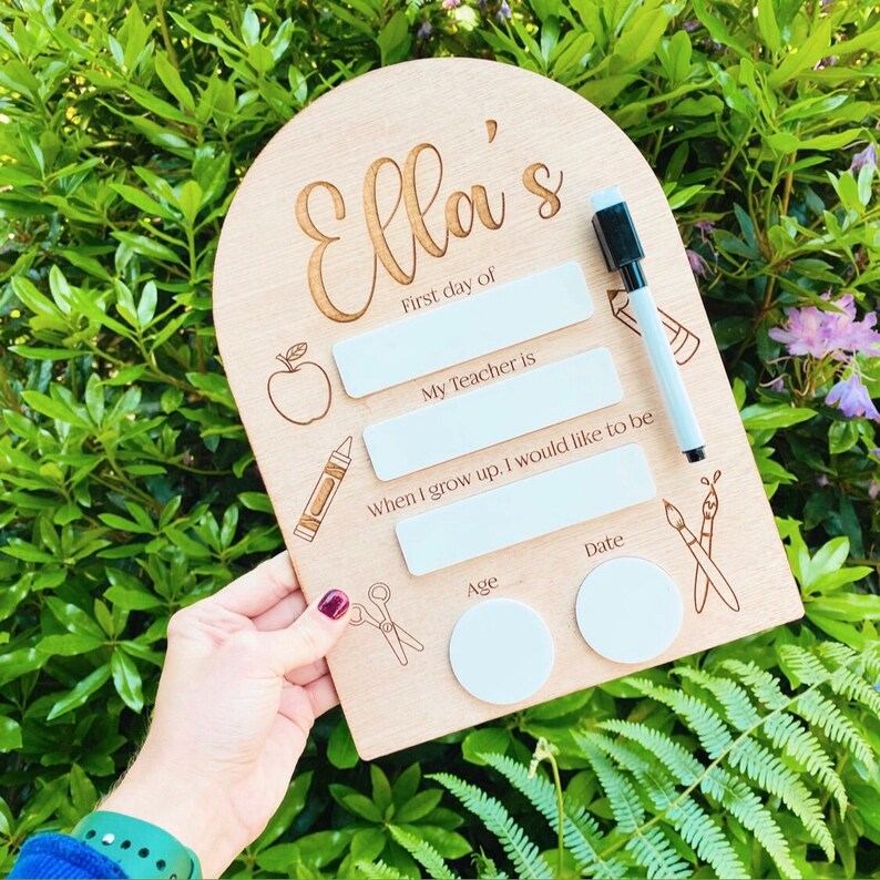 May include: A wooden sign with the name "Ella's" engraved on it. The sign has blank spaces for writing with a dry-erase marker, including "First day of", "My Teacher is", "When I grow up, I would like to be", "Age", and "Date".