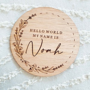 May include: A circular wooden plaque engraved with the words "HELLO WORLD MY NAME IS Noah". The text is in black script, surrounded by a floral wreath. The wood has a natural grain.