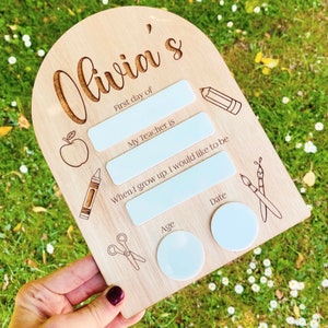 May include: A wooden sign with the text "Olivia's" and spaces for a child's first day of school information. The sign has a cutout of an apple, a crayon, scissors, and paintbrushes.