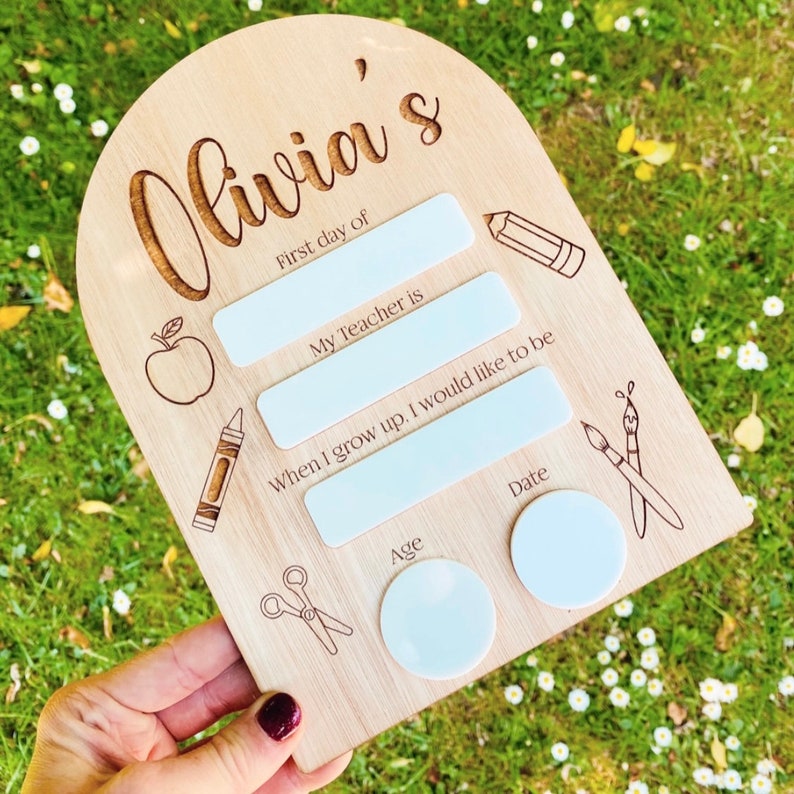 May include: A wooden sign with the text "Olivia's" and spaces for a child's first day of school information. The sign has a cutout of an apple, a crayon, scissors, and paintbrushes.