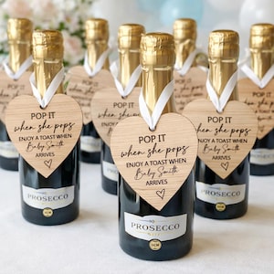 Puede incluir: Una colección de mini botellas de Prosecco, cada una con una etiqueta de madera clara en forma de corazón atada con una cinta blanca. Las etiquetas tienen impreso el texto: POP IT when she pops ENJOY A TOAST WHEN Baby Smith ARRIVES, con un corazón.
