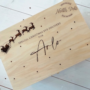 Puede incluir: Una caja de madera con un tema de entrega de Nochebuena. La caja está decorada con una silueta de Papá Noel y sus renos volando por el aire. El texto "SPECIAL CHRISTMAS EVE DELIVERY FOR Arlo" está grabado en la caja. La caja también tiene un pequeño diseño de estrella.