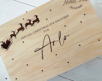 Op de afbeelding: Een houten doos met een kerstmis-avondleveringsthema. De doos is versierd met een silhouet van Sinterklaas en zijn rendieren die door de lucht vliegen. De tekst "SPECIAL CHRISTMAS EVE DELIVERY FOR Arlo" is in de doos gegraveerd. De doos heeft ook een klein sterrenthema.