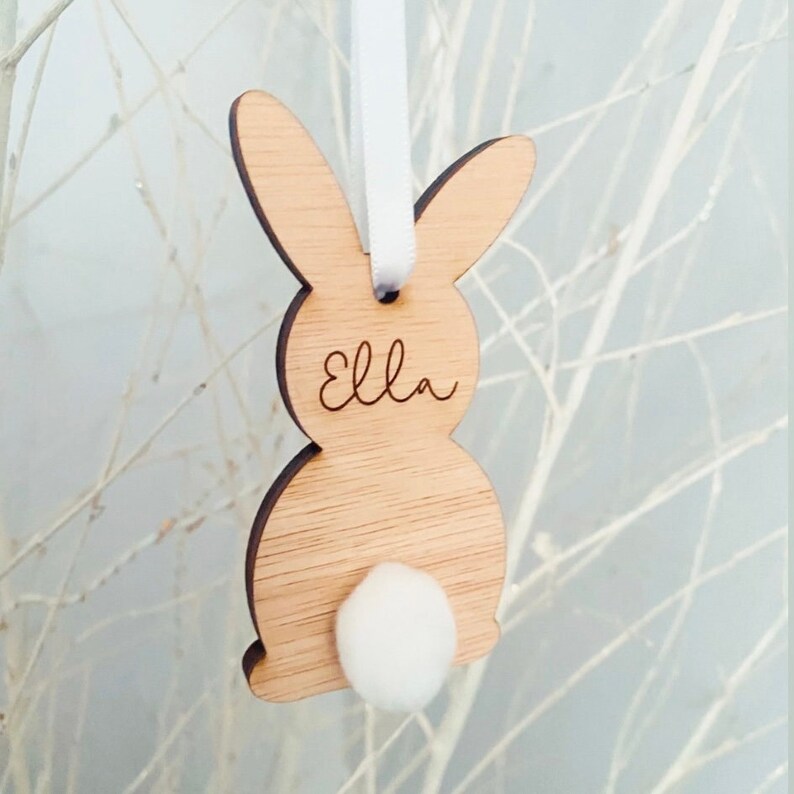 May include: A wooden bunny rabbit ornament with a white fluffy tail. The bunny is light brown and has the name "Ella" engraved on its chest.