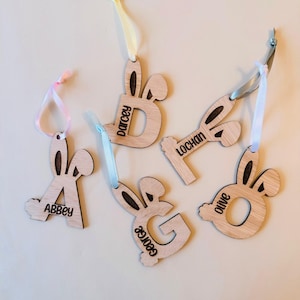 May include: A collection of wooden Easter bunny-shaped ornaments, each featuring a letter and a name. The letters are "A", "B", "G", "L", and "O", with names "Abbey", "Darcey", "George", "Lochan", and "Olive". Each has a ribbon for hanging.