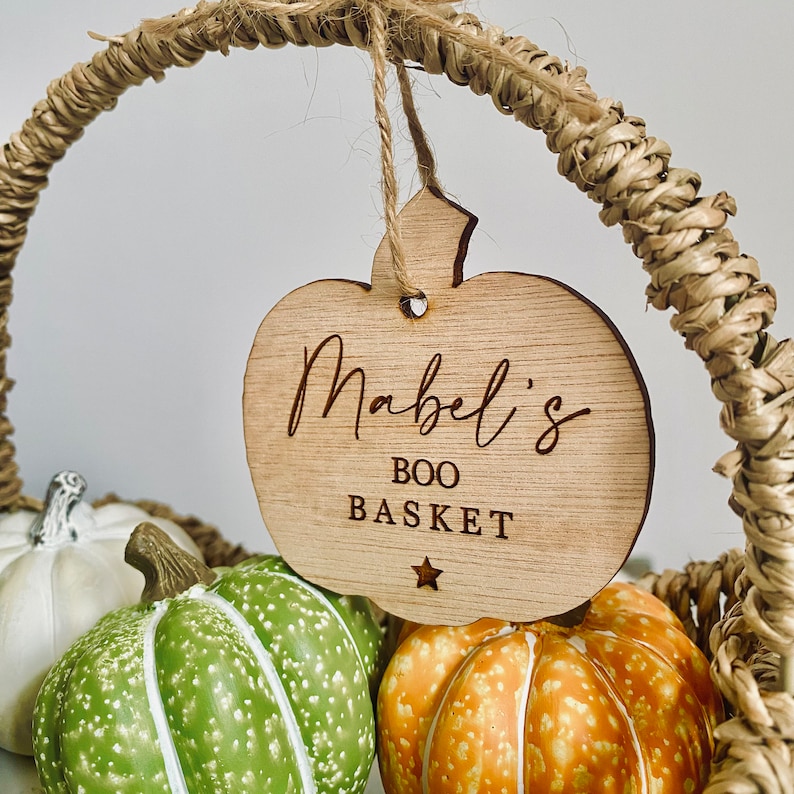 Puede incluir: Una etiqueta de madera con forma de calabaza con el texto "Mabel's Boo Basket" y una peque&ntilde;a estrella. La etiqueta est&aacute; colgada de una cesta de mimbre llena de peque&ntilde;as calabazas.