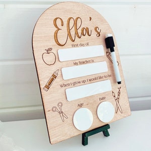 May include: A wooden sign with the name "Ella's" engraved on it. The sign has blank spaces for writing information about the first day of school, including the child's teacher, what they want to be when they grow up, their age, and the date.
