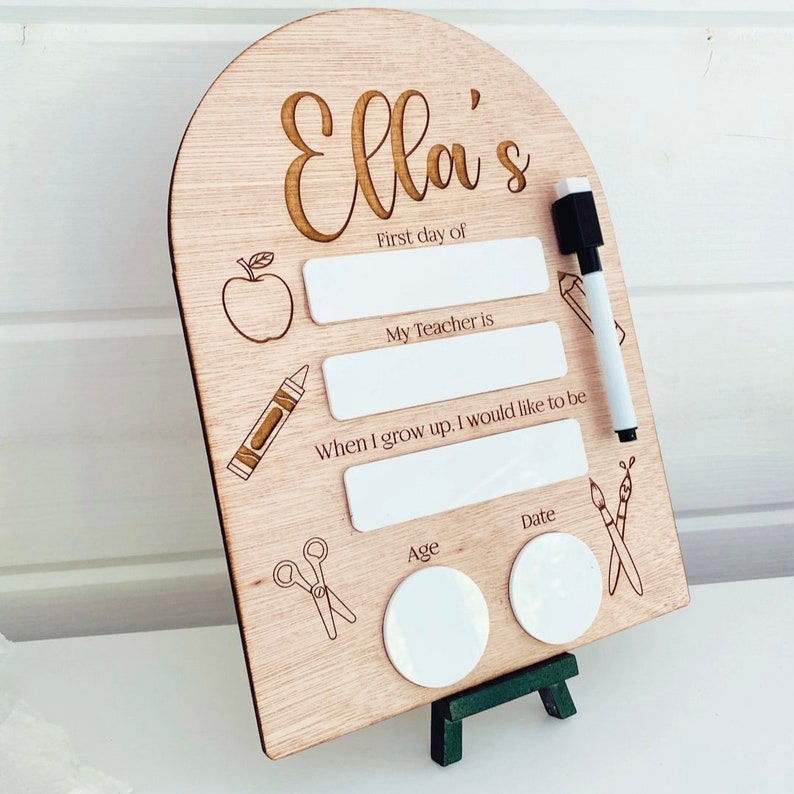 May include: A wooden sign with the name "Ella's" engraved on it. The sign has blank spaces for writing information about the first day of school, including the child's teacher, what they want to be when they grow up, their age, and the date.