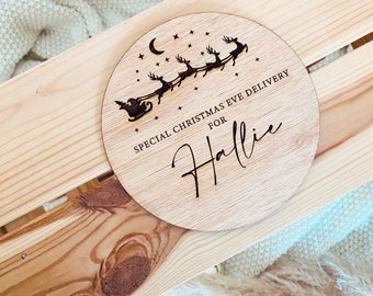 Pode incluir: Um sinal redondo de madeira com um Papai Noel e renas voando pelo ar com uma lua crescente e estrelas no fundo. O sinal diz "Entrega especial de véspera de Natal para Hallie".