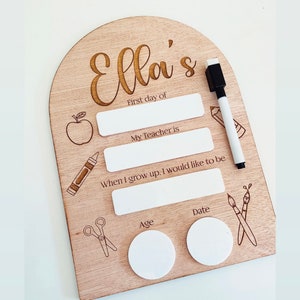 May include: A wooden sign with the name "Ella's" engraved on it. The sign has spaces for writing the first day of school, the teacher's name, what the child wants to be when they grow up, their age, and the date. The sign is decorated with drawings of an apple, a crayon, scissors, and paintbrushes.