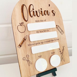 May include: Wooden sign with the name "Olivia's" engraved on it. The sign has spaces to fill in the first day of school, teacher's name, future career, age, and date. The sign is decorated with drawings of an apple, crayons, scissors, and paintbrushes.