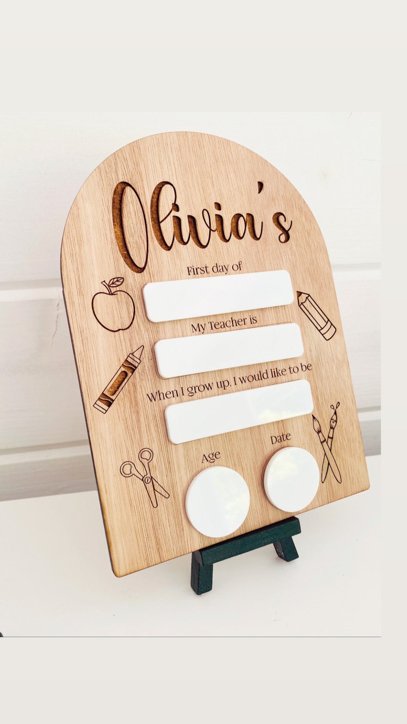 May include: Wooden sign with the name "Olivia's" engraved on it. The sign has spaces to fill in the first day of school, teacher's name, future career, age, and date. The sign is decorated with drawings of an apple, crayons, scissors, and paintbrushes.