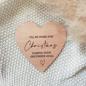 May include: A wooden heart-shaped sign with the text "I'll be home for Christmas coming soon -December 2025-." The sign is on a white knitted surface.