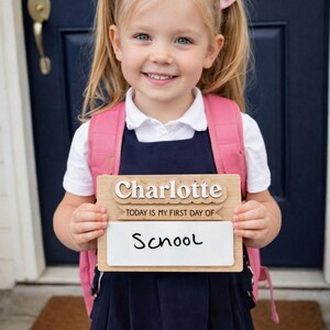 May include: A young girl with blonde hair and pink bows holds a wooden sign that says "Charlotte" and "Today is my first day of School". She wears a pink backpack, a white shirt, and a navy blue dress.