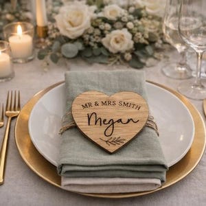 May include: A wooden heart-shaped place card with the text "MR & MRS SMITH" and the name "Megan" in cursive script. The place card rests on a folded green napkin, atop a white plate with a gold rim, set for a wedding.