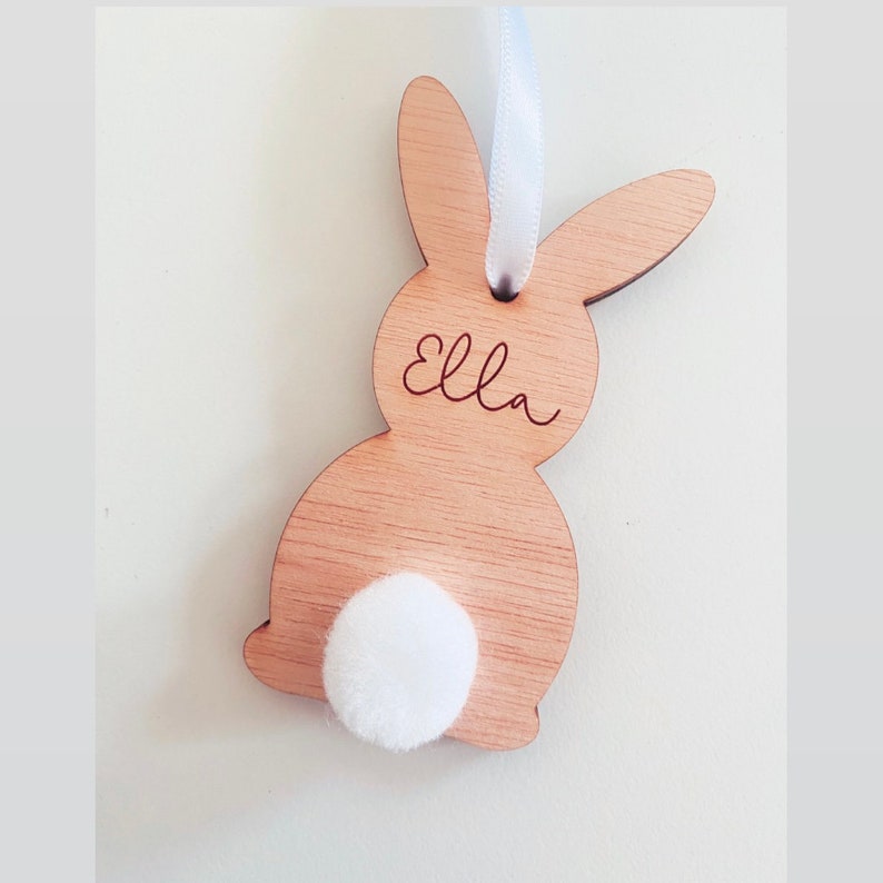 May include: A wooden bunny rabbit ornament with a white pom-pom tail and the name "Ella" engraved on the front.