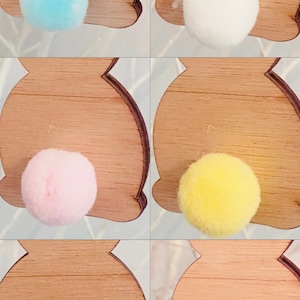 May include: Five wooden bunny shapes with different colored pom-poms attached to the back. The colors are light blue, white, pink, yellow, and brown.