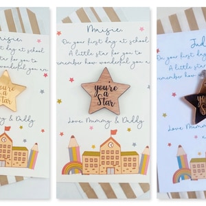 May include: Three cards with a star-shaped wooden charm attached. Each card has a different name on it: Maisie, Maisie, and Jude. The cards have a message about the first day of school and a schoolhouse illustration. The star charms have the words "you're a star" engraved on them.