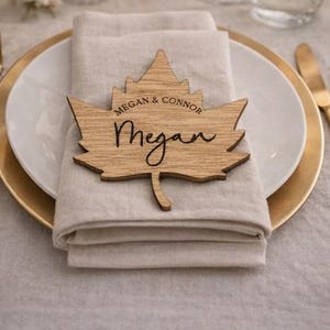 May include: A wooden maple leaf place card with the name "Megan" in cursive script. The place card also includes the names "Megan & Connor" in a smaller font. The place card is placed on a folded beige napkin, atop a white plate with a gold rim.