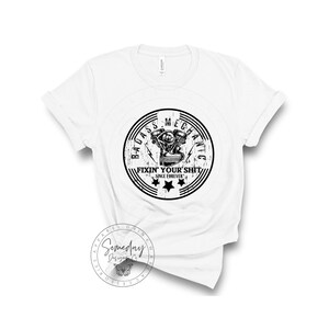 May include: White t-shirt with a black and white graphic of a motorcycle engine in a circle with the text "Badass Mechanic Fixin' Your Shit Since Forever".