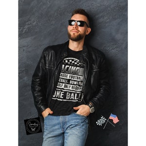 Op de afbeelding: Een man draagt een zwart T-shirt met een witte afbeelding waarop staat "Racing because football, baseball, bowling, golf only requires one ball". Hij draagt een zwart leren jack en een blauwe jeans. Hij staat voor een grijze muur.