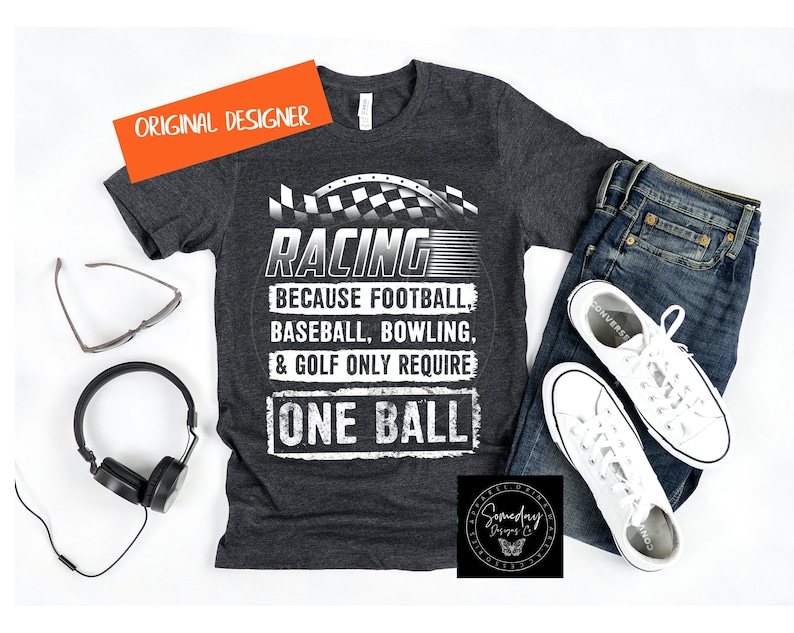 Op de afbeelding: Een donkergrijs T-shirt met een witte afbeelding waarop staat "Racing because football, baseball, bowling, & golf only require one ball". De afbeelding bevat ook een geruit vlagontwerp.