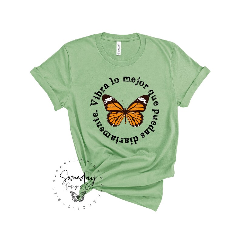 K&ouml;nnte beinhalten: Ein hellgr&uuml;nes T-Shirt mit einer runden Grafik, die einen Schmetterling und den Text "Vibra lo mejor que puedas diariamente." zeigt.