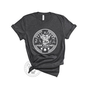 May include: A dark gray t-shirt with a white distressed circle graphic that says "Badass Mechanic Fixin' Your Shit Since Forever" with a stylized engine illustration inside the circle.