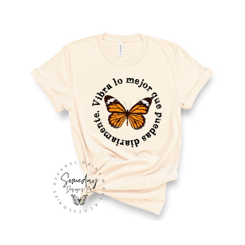K&ouml;nnte beinhalten: Ein beiges T-Shirt mit einem schwarzen Kreis-Motiv, das einen Schmetterling und den Text "Vibra lo mejor que puedas diariamente." zeigt.