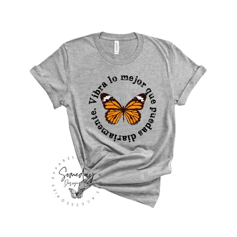 K&ouml;nnte beinhalten: Graues T-Shirt mit einem schwarzen Kreis-Motiv, das einen Schmetterling und den Text "Vibra lo mejor que puedas diariamente." zeigt.