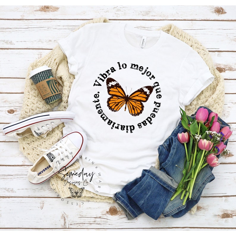 K&ouml;nnte beinhalten: Wei&szlig;es T-Shirt mit einem schwarzen Kreis-Motiv, das einen Schmetterling und den Text "Vibra lo mejor que puedas diariamente." zeigt.