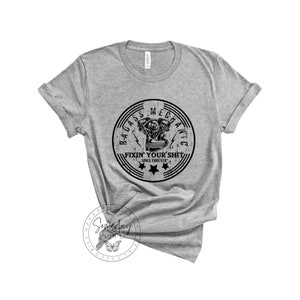 May include: Gray t-shirt with a black and white graphic print of a motorcycle engine. The graphic has the text "Badass Mechanic Fixin' Your Shit Since Forever".