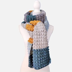 Op de afbeelding: Een gebreide sjaal met een veelkleurig patroon in tinten blauw, grijs en geel. De sjaal wordt getoond op een wit mannequin.