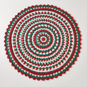 Op de afbeelding: Een rond, gehaakt tafellaken in rood, wit en groen. Het tafellaken heeft een stervormig midden en meerdere lagen concentrische cirkels.