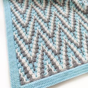 Op de afbeelding: Een blauwe, witte en grijze gehaakte babydeken met een chevron-patroon. De deken heeft een blauwe rand.