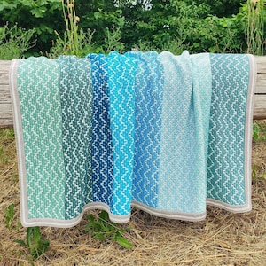 Op de afbeelding: Een gehaakt deken met een gradiënt van blauwe en groene kleuren. Het deken heeft een witte rand en een grijze franje.