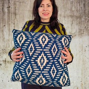 Op de afbeelding: Een vrouw houdt een blauw en wit gepatroneerd kussen vast met een geometrisch ontwerp. Het kussen is gemaakt van een zachte, textuurrijke stof.