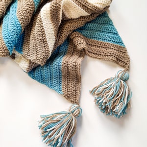 Op de afbeelding: Een gestreept gebreid deken met kwastjes in bruin, wit en blauw.